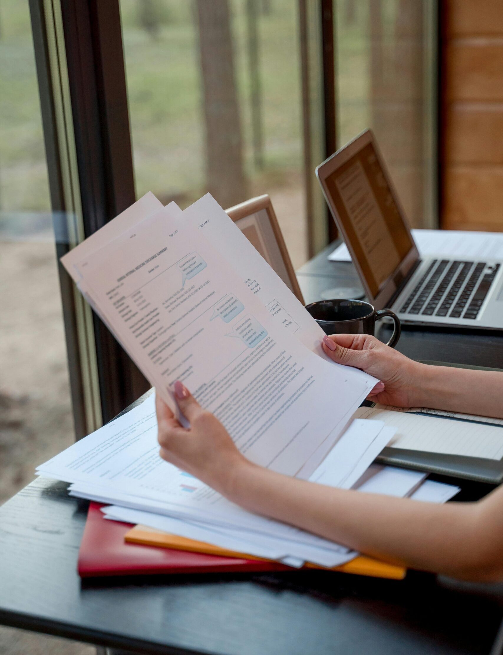 Person holding and reviewing documents near window with laptops on a desk.